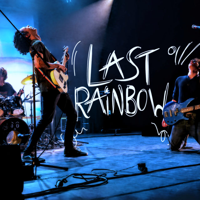 Image du site du groupe LAST RAINBOW prise lors d'un concert à Cannes au Théâtre de la Licorne. Le guitariste et le bassiste se font face et le logo de LAST RAINBOW est dessiné entre eux deux.