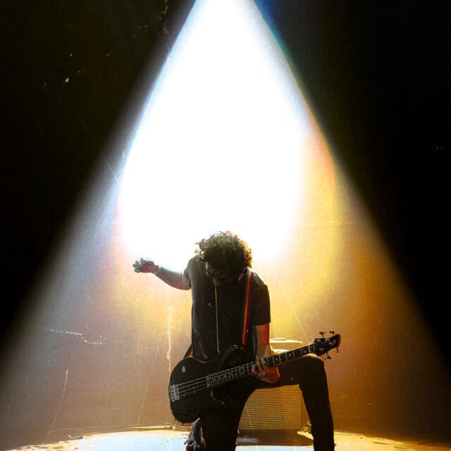 Kénan Saporito le bassiste de LAST RAINBOW prend la pose lors d'un concert au Stockfish. Il pose un genou au sol, un spot lumineux l'éclair depuis le haut de la salle