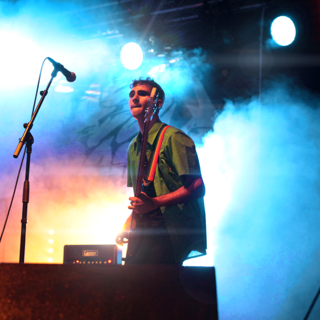 Kénan Saporito le bassiste de LAST RAINBOW joue de la basse lors d'un concert pour la fête de la musique à Aix-en-Provence organisé par Class'eurock. Le bassiste est entouré de fumée bleue et jaune ce qui fait ressortir sa veste verte et son grand nez