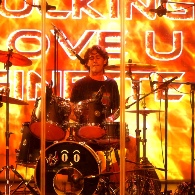 Raphaël Meyer le batteur de LAST RAINBOW lors d'un concert à Cannes au théâtre de la licorne. Il joue de la batterie derrière une vitre en plexiglas. Derrière, un écran affiche des flammes énormes qui colorent toute la scène de couleurs chaudes