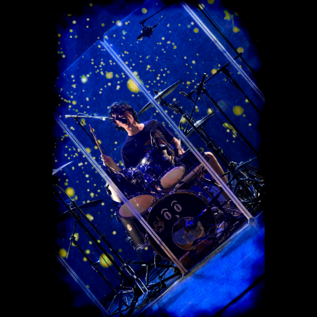 Raphaël Meyer le batteur de LAST RAINBOW lors d'un concert à Cannes au théâtre de la licorne. Il joue de la batterie derrière une vitre en plexiglas. Derrière lui, un écran affiche un fond bleu et des particules jaunes faisant ressembler l'image à un tableau de Van Gogh