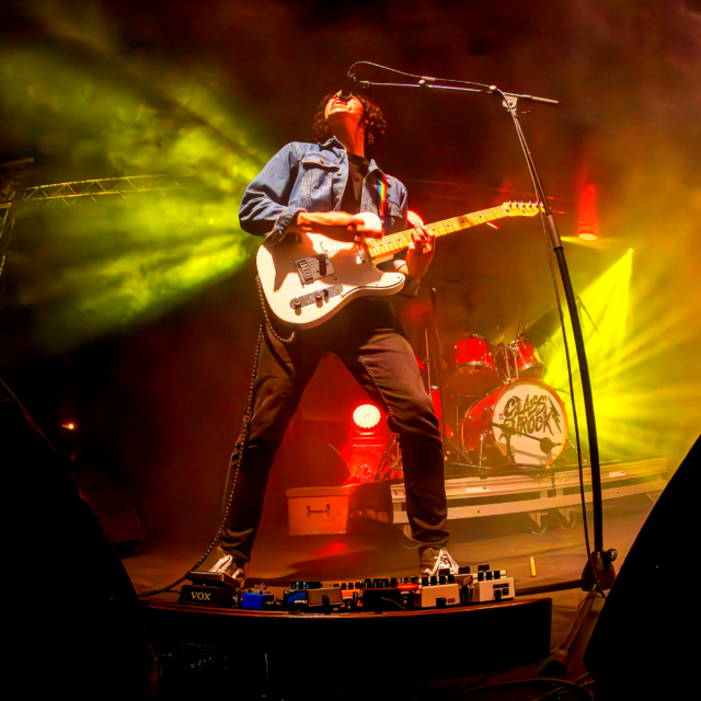 Alex Hutin le guitariste chanteur de LAST RAINBOW joue lors de la fête de la musique 2024 à Aix-en-Provence organisée par Class'eurock. La photo est en contre plongée, et la veste bleue d'Alex ressort en contraste avec les projecteurs jaunes et rouges derrière lui